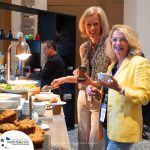 Two women smiling and holding coffee cups at a buffet table with muffins and various dishes. A man in the background is serving himself. The setting appears to be a professional event or conference.