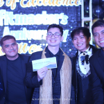 Five men stand together at an event, smiling for a group photo. One man holds an envelope and a certificate. The background displays text and a balloon decoration.