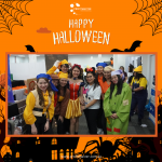 People in Halloween costumes smile for a group photo at an indoor event. The frame has a Halloween-themed border.