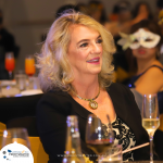 A woman with blonde hair sits at a formal event, wearing a black top and necklace. She is smiling. Other guests are in the background, some wearing masks. Wine glasses are on the table.