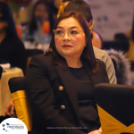 A woman with glasses and a black blazer sits at a formal event. She appears attentive, with a neutral expression. The background shows other attendees and event decorations.