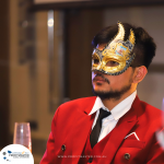 A man in a red suit jacket and a decorative mask with gold and blue accents sits in an indoor setting, next to a champagne glass. The backdrop includes a logo reading "Profitmaster.