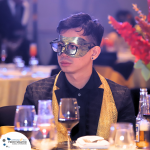 A person in formal attire wears a decorative mask at a table set with drinks and glassware. Brightly colored flowers are in the background.