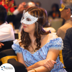 A woman in a blue and white dress wearing a white masquerade mask sits at an event.