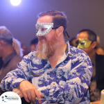 A man with a long beard, wearing a decorative mask and a blue patterned shirt, seated among others at an event.