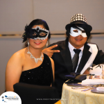 Two people wearing formal attire and masquerade masks sit at a table; one gives a peace sign. A pamphlet and some items are on the table, with a logo and website at the bottom left corner.