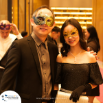 A man and woman in formal attire wearing decorative masks stand side by side at a masked event. Other masked attendees are visible in the background.