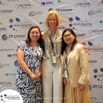 Three women pose for a photo at a business event, standing in front of a branded backdrop featuring multiple company logos. They are smiling and wearing name badges.