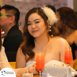 A woman in an off-shoulder top with a feather hair accessory sits at a table with two orange-pink drinks in front of her. Other people are in the background.