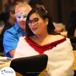 A woman in a red dress and white fur shawl sits at a formal event. A person wearing a blue shirt and a masquerade mask is visible in the background.