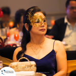 A woman in a blue dress and a gold mask sits at a table adorned with a purse. Other attendees can be seen in the background at the formal event.