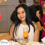 A woman with long black hair smiles at the camera while seated at a table. A drink in a tall glass is in front of her. Other people are in the background of the indoor setting.