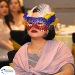 A person wearing a decorative mask with red, blue, and yellow accents and feathers, dressed in formal attire at an indoor event.