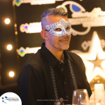 A person wearing a silver masquerade mask and a black shirt sits at a table with blurred festive lights in the background.