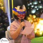 Person wearing a colorful masquerade mask and a lavender shawl is speaking into a microphone while holding a sheet of paper. The background features balloons and festive decorations.