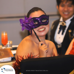 Person wearing a purple mask with a flower, smiling at a formal event. Another person in formal attire is in the background. Books and a drink are on the table.