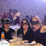 Three people wearing masquerade masks are seated at a table during an event, smiling at the camera. The background features a step and repeat banner with logos.