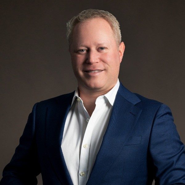 A man with short, light hair wearing a dark blue suit jacket and white shirt stands in front of a dark background, smiling at the camera, likely celebrating the 10th Anniversary of Profitmaster at a conference.