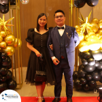 A man in a blue suit and a woman in a black dress pose together on a red carpet, surrounded by black and gold balloons.