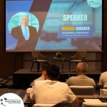 A conference room with a large screen displaying a slide introducing Richard Croaker, Founder and President of Profitmaster Global Outsourcing, as a speaker on "Building a Global Workforce." Attendees are seated.