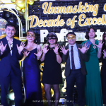 Eight people standing in a row wearing formal attire and masks, posing with raised hands before a backdrop that says "Unmasking a Decade of Excellence: Refined Power." Decor includes balloons and lights.