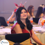 A woman in a red dress sits at a decorated table during an event. She has dark hair and wears a floral headpiece. The background shows other guests and tables.