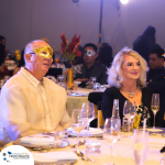 A man and a woman, both dressed elegantly, are seated at a dining table during a formal event. The man is wearing a gold masquerade mask. The table is set with glasses and plates.