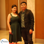 A man and a woman posing together in front of a wooden paneled wall. The woman is wearing a green dress and holding a phone, while the man is dressed in a suit jacket and shirt.