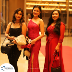 Three women dressed in formal attire stand on a red carpet in a brightly lit venue.