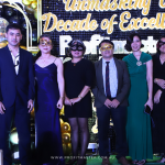 A group of people in formal attire and masquerade masks pose in front of a backdrop reading "Celebrating a Decade of Excellence.”.