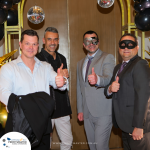 Four men, two in masks, dressed in formal attire give thumbs up at an event. The backdrop features balloons, disco balls, and an archway. A sign reads "Profitmaster Group".