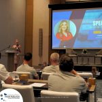 A speaker stands at a podium addressing an audience in a conference room. A presentation is displayed on a screen behind her, showing the title "Speaker: Marketing Strategies" along with a photo and text.