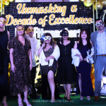 A group of people wearing masquerade masks pose in front of a backdrop with the text "Unmasking a Decade of Excellence." Balloons and decorations surround them.