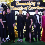 A group of nine people stands in formal attire in front of a backdrop that reads "Unmasking a Decade of Excellence" with golden decorations and balloons.