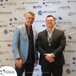 Two men in business attire stand in front of a branded backdrop at a professional event.