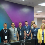 Group of eleven people standing in an office room, posing for a photo. Several individuals wear ID badges. A banner with the logo "ProfitMaster" is visible in the bottom left corner.