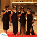 Four women wearing elegant dresses and masquerade masks stand on a red carpet in a formal setting.