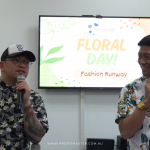 Two men in floral shirts speak into microphones in front of a screen displaying "Floral Day! Fashion Runway.