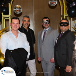 Four men, two wearing masquerade masks, pose in front of a decorated backdrop with balloons and hanging glitter balls. A sign in the bottom left corner reads "Profitmaster.