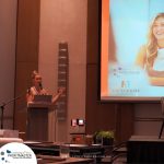 A woman stands at a podium speaking into a microphone in a conference room. A large screen beside her displays a promotional image for Profitmaster Global Outsourcing.