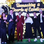 A group of people in formal attire standing in front of a gold and black-themed backdrop that reads "Unmasking a Decade of Excellence: Profit Master". Most are wearing decorative masks.