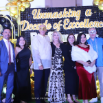 A group of nine people, some wearing masks, stands in front of a "Unmasking a Decade of Excellence" backdrop with gold and black balloons.