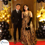 Two individuals in formal attire stand on a red carpet. The background features star decorations and balloons.