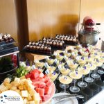 A buffet table featuring various desserts including chocolate cakes, custards in glasses topped with flowers, and a tray of cut fruits like watermelon and pineapple. An electric mixer is in the background.