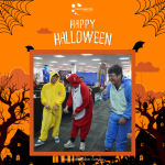 Three people in colorful animal onesies laughing in an office decorated for Halloween with spider webs and bats.