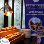 A table with plated food, illuminated by a lamp. In the background, a banner reads "10th Anniversary Conference 2021 ProfitMaster. Welcome speakers and delegates." Plates are stacked on the table.