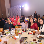 A group of people dressed formally, seated around a table at a banquet. The table has drinks, plates of food, and floral centerpieces. Some people are smiling and one person is wearing a mask.