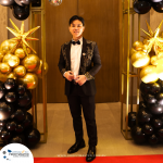 A person dressed in formal attire stands on a red carpet between two balloon columns decorated with black and gold balloons and gold star-shaped balloons.