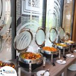 A buffet setup with multiple chafing dishes containing various food items. Each dish has a lid partially open, and plates are placed beside the dishes. A logo and website are visible on the image.