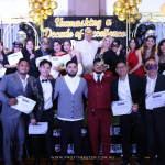 A group of people dressed in formal wear and masks pose for a photo at an event. Some hold certificates. Behind them is a sign that reads, "Unmasking a Decade of Excellence.